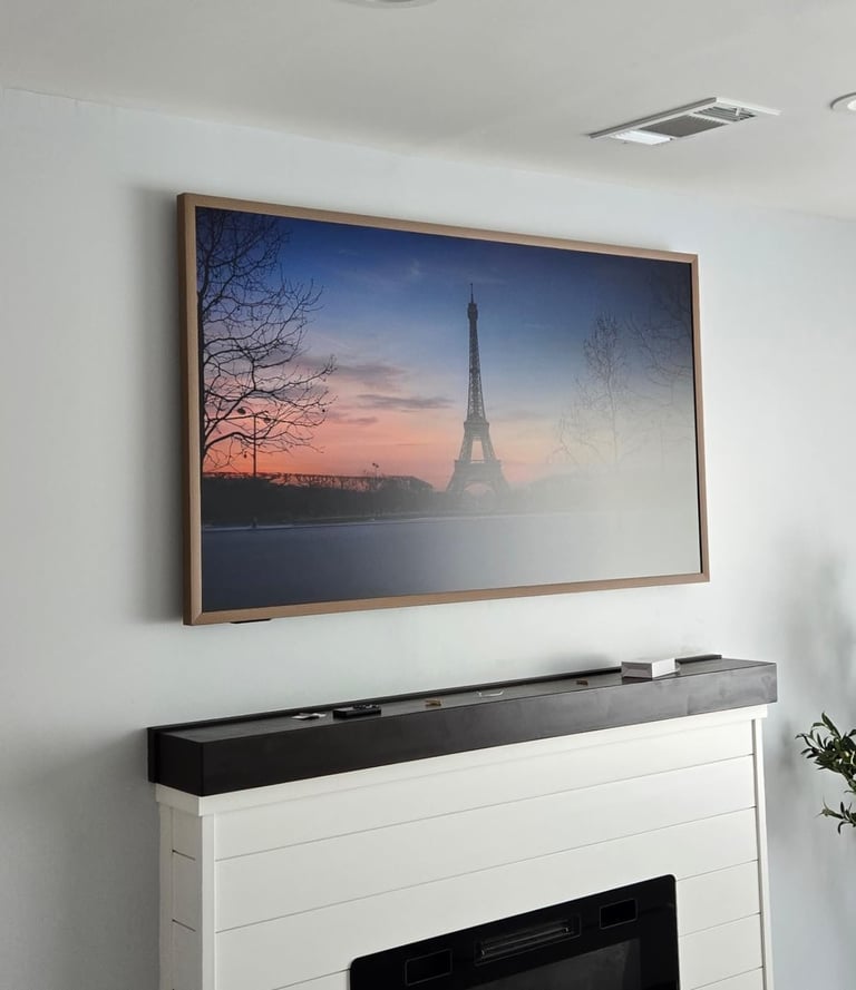 Framed TV on white wall above fireplace displaying Eiffel Tower at sunset with bare trees and water reflection