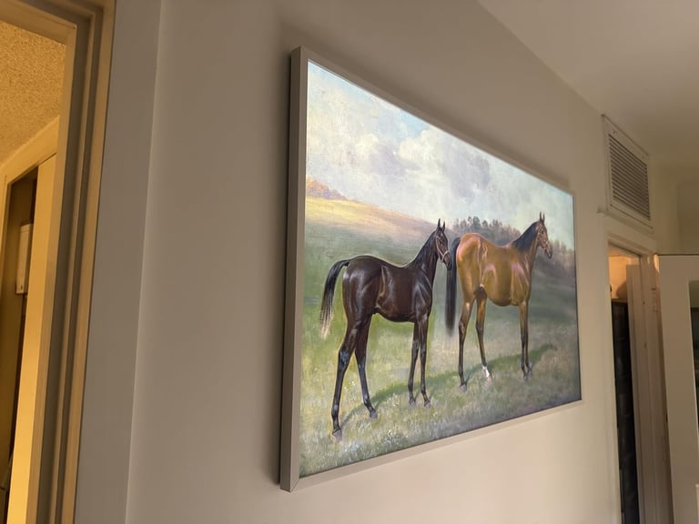 Framed painting of three horses standing in a pastoral landscape, mounted on a beige wall between two doorways
