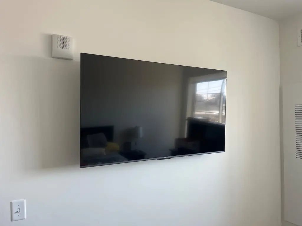 Flat-screen TV mounted on a plain cream wall with a window reflected in the screen